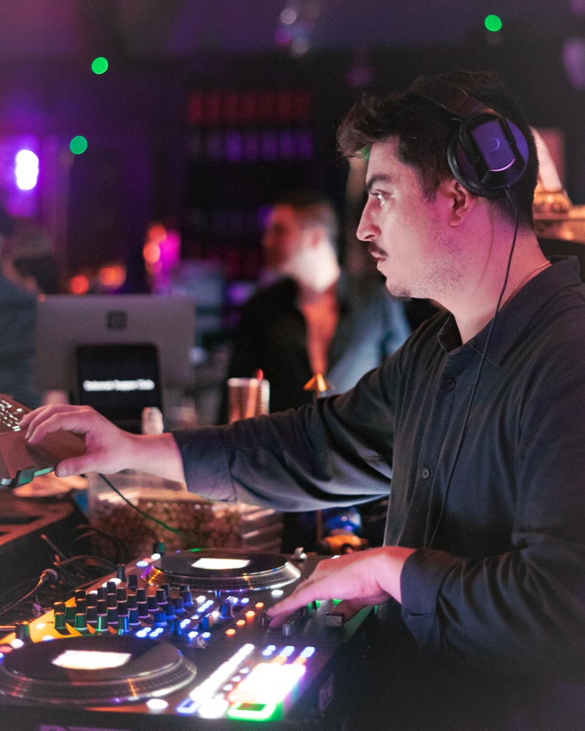 DJ OTEO sharp profile at the rotary mixer, club-glow bokeh, working hands on jogwheel