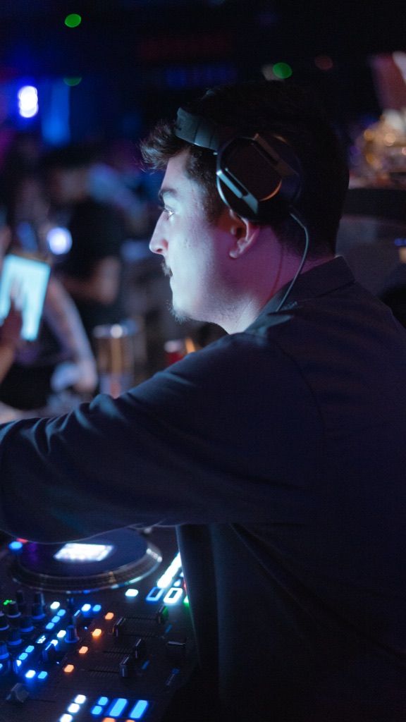 DJ OTEO profile at the deck mid-mix, blue + amber stage-light bokeh