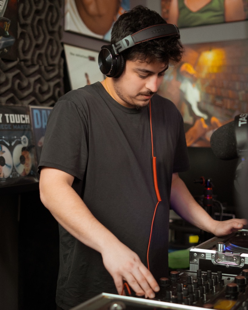 DJ OTEO at the decks, headphones on, hand on the mixer, eyes-down working